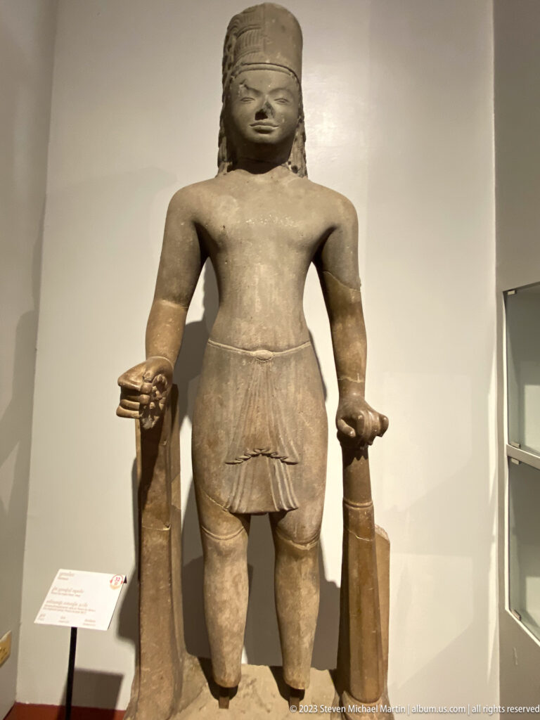 Standing sandstone figure with carved side supports displayed on a museum pedestal.