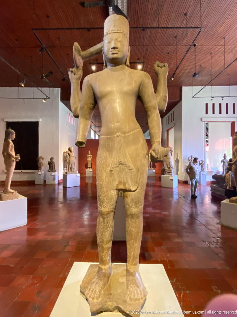 Standing four-armed sandstone deity displayed in the National Museum of Cambodia gallery.