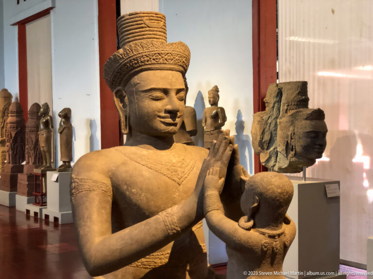 Standing crowned figure holding a ritual vessel displayed in the National Museum of Cambodia gallery.