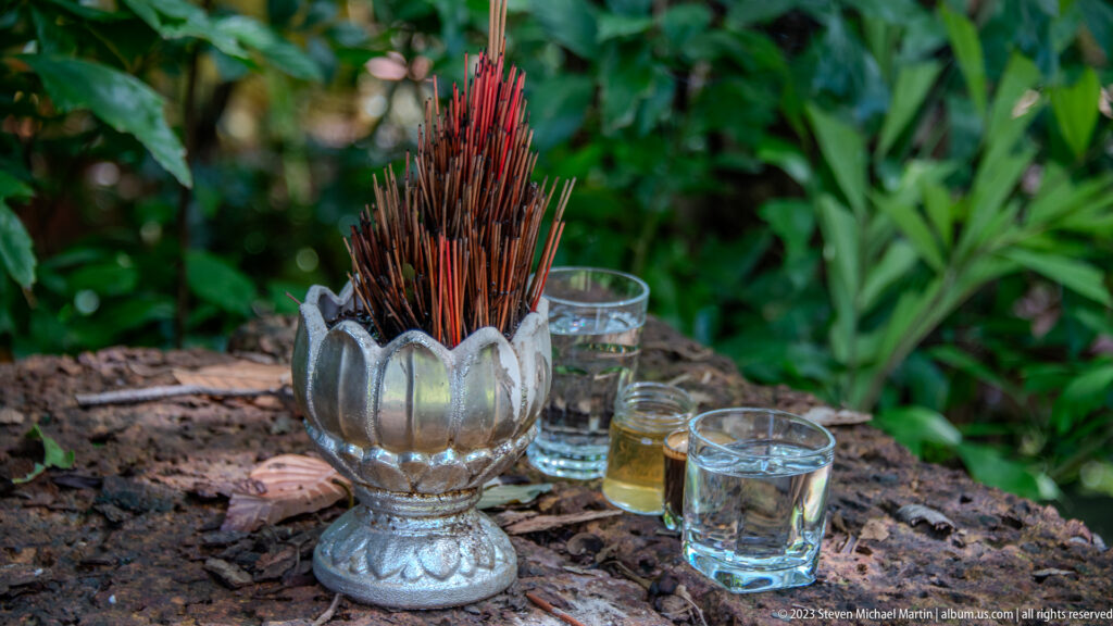 Red incense sticks in a silver lotus-shaped holder surrounded by small glasses of clear and amber liquid.