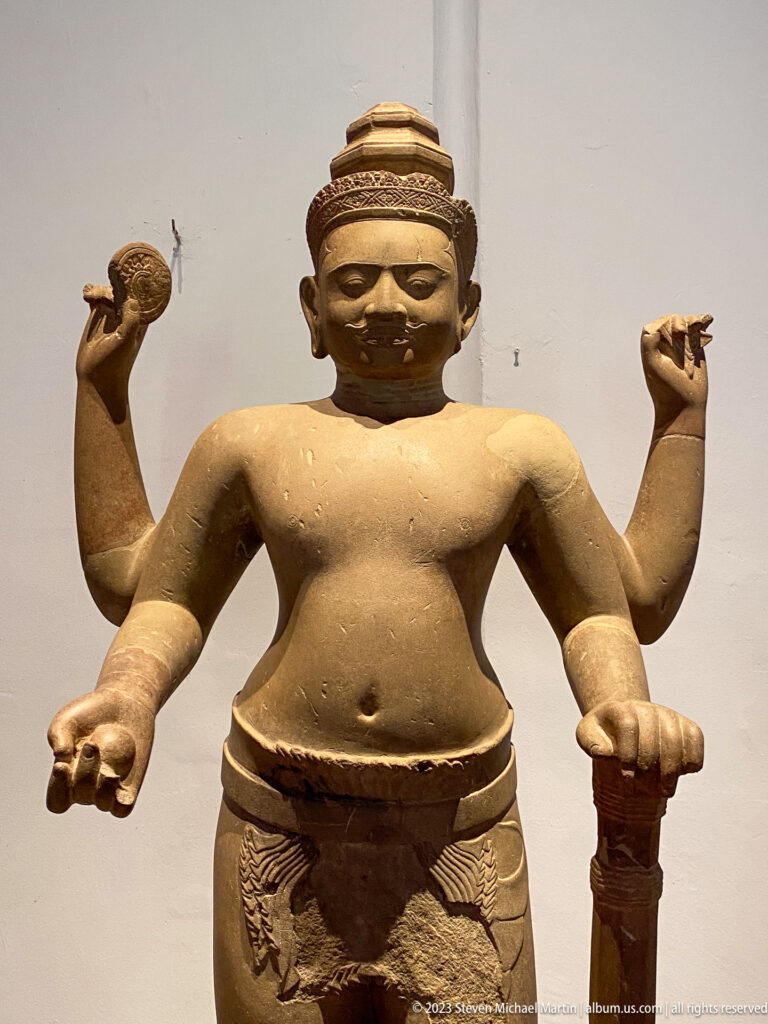 Standing four-armed sandstone deity displayed in the National Museum of Cambodia gallery.