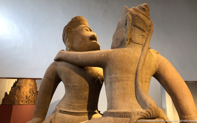 Two interacting sandstone figures displayed in the National Museum of Cambodia gallery.