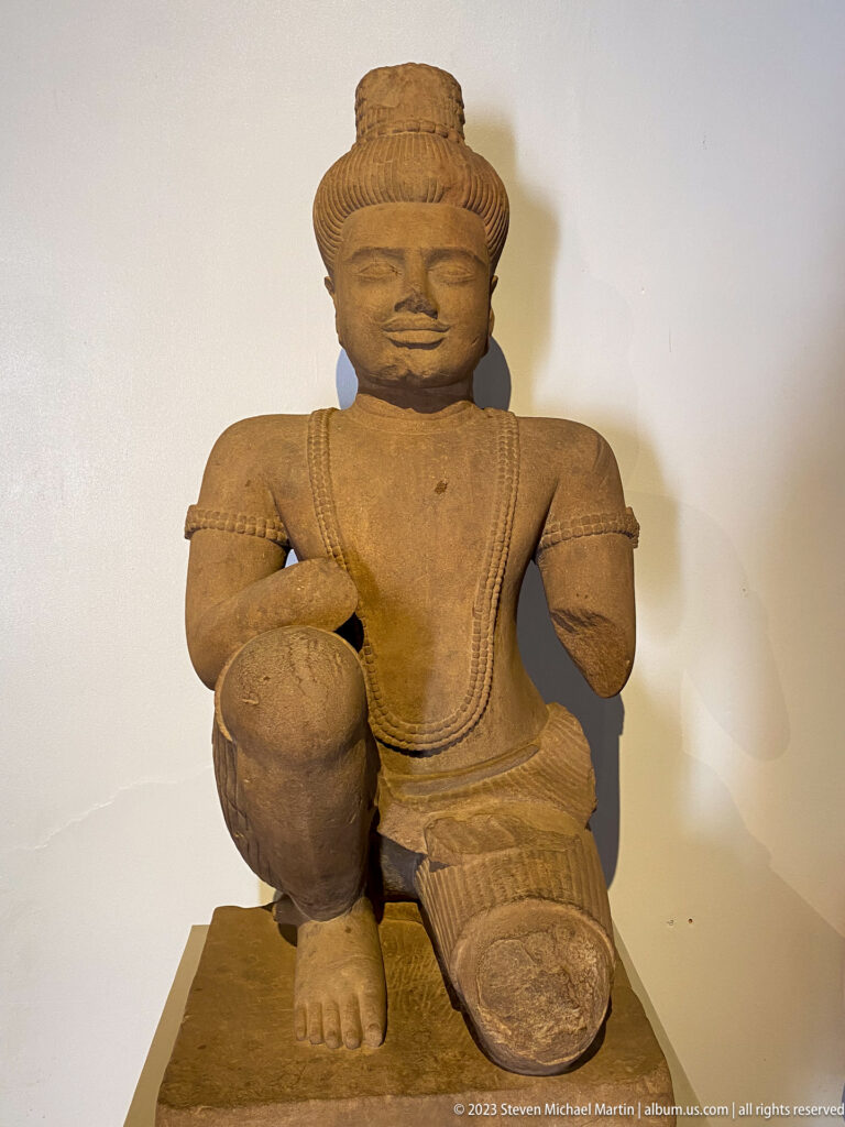 Kneeling sandstone figure with one raised knee, jewelry, drapery, and damaged arms.