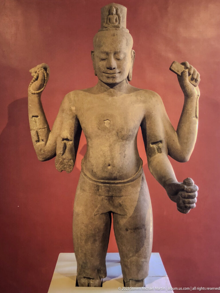 Standing four-armed sandstone deity with tall headdress against a deep red gallery wall.