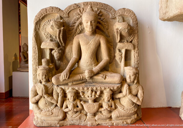 Seated Buddha beneath a multi-headed naga canopy with kneeling attendants and carved foliage.