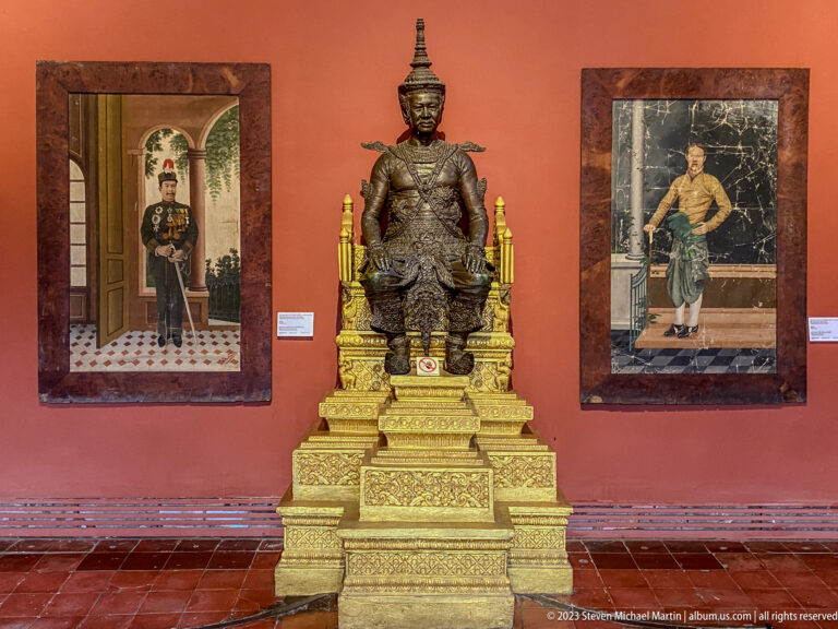 Seated royal figure on an elaborate gilded throne between formal portraits on a red wall.