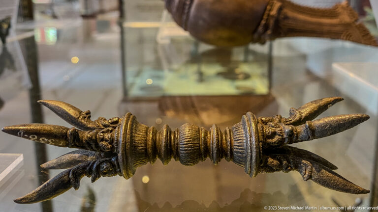 Double-ended bronze vajra displayed horizontally inside a protective glass museum display case.