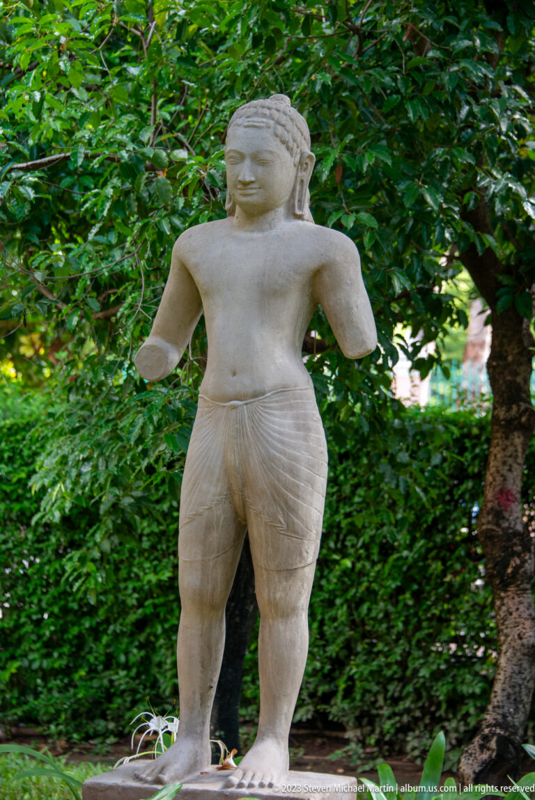 Weathered stone guardian with broad shoulders, bent arms, and powerful legs standing against dense green foliage.