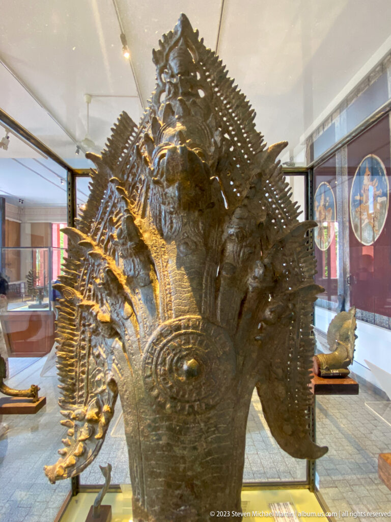 Large bronze mythological guardian surrounded by elaborate radiating ornament inside a display case.