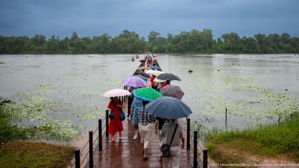 Umbrella-carrying visitors walk along a wet boardwalk through lotus-covered water beneath a dark sky.