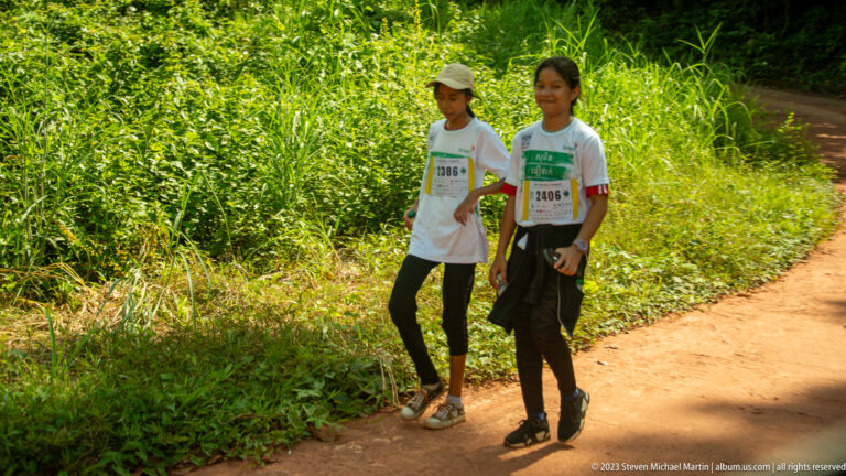 Two young women in numbered white shirts walk on a dirt path through dense green vegetation.