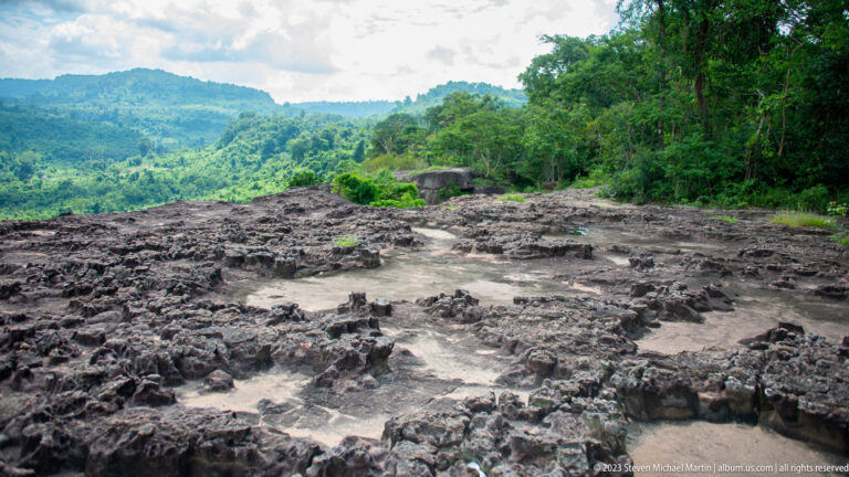 Low stone wall foundations and rubble exposed on a rocky clearing beside dense forest.