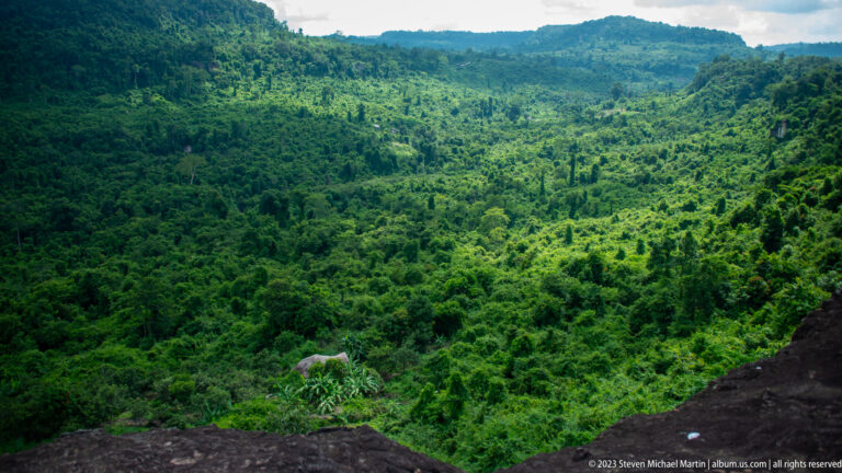 Dense green forest valley and layered ridges viewed from the edge of a dark rock ledge.