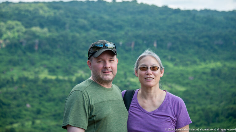 Two adult visitors pose together with green forested hills softly blurred behind them.