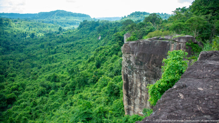 Angular rock promontory juts over a lush forest valley with green ridges in the distance.