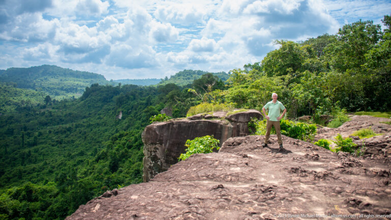Man stands on a rocky cliff overlook above dense forest and distant green ridges.