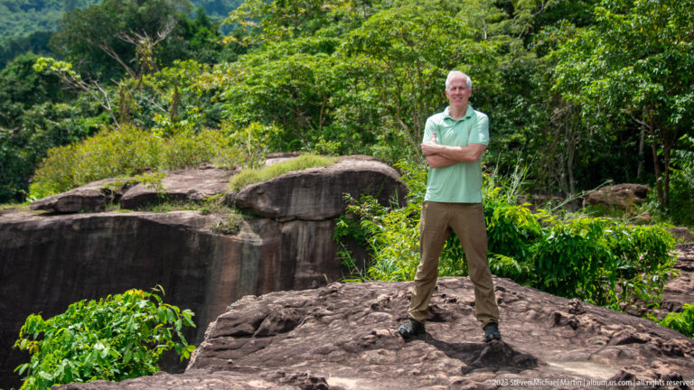 Older man in a green shirt stands with folded arms on rock beside a forested cliff.