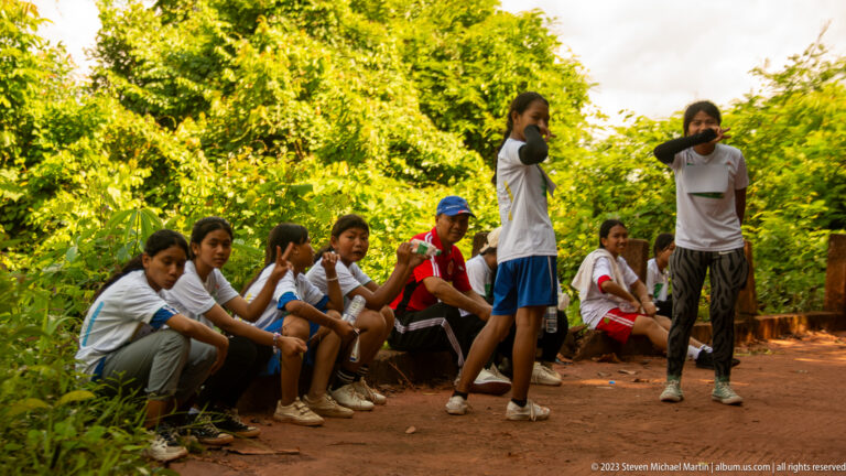 Young participants in matching shirts rest and stand beside a shaded dirt road in the forest.