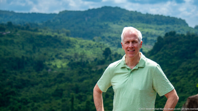 Smiling older man in a pale green shirt with blurred forested hills behind him.
