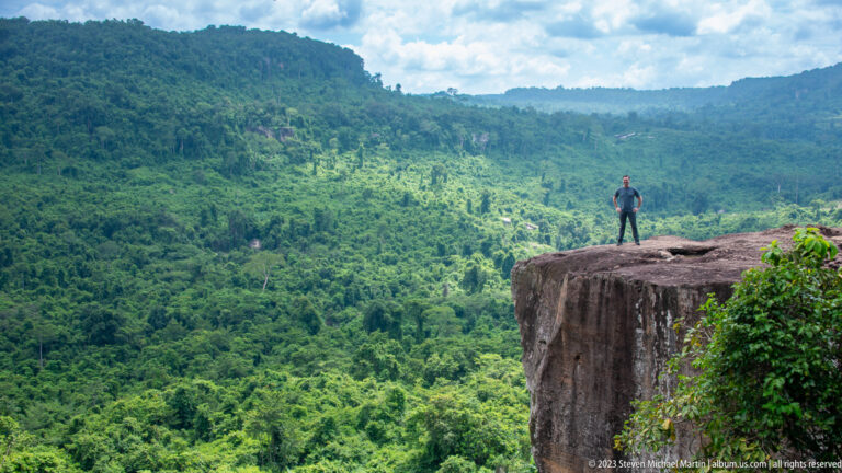 Small lone figure stands on a high rock promontory above a vast green forest valley.