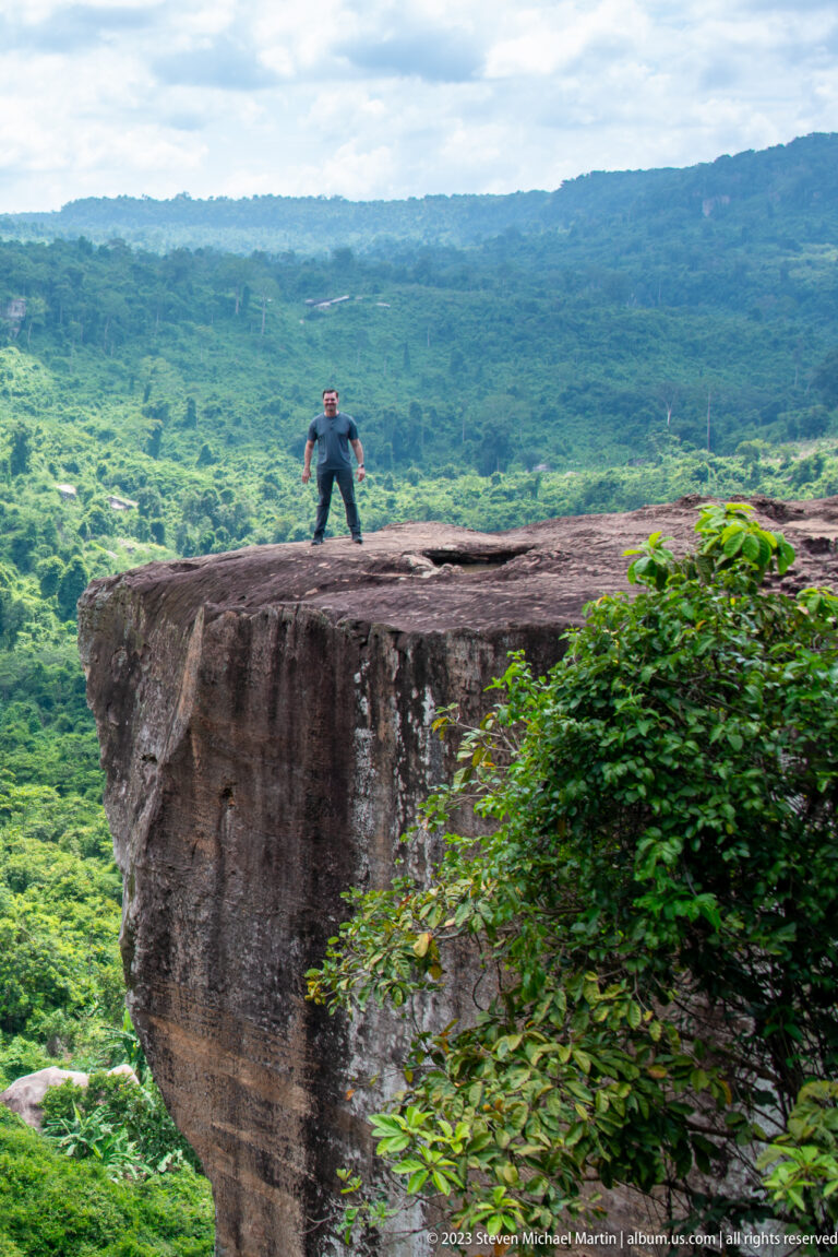 Visitor crouches on a wide flat cliff ledge above an expansive green forested landscape.