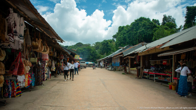 Visitors walk along a market lane lined with open stalls selling textiles, bags, and souvenirs.