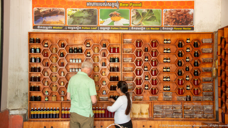 Honey bottles and bee products displayed in hexagonal shelves while a customer speaks with a shopkeeper.