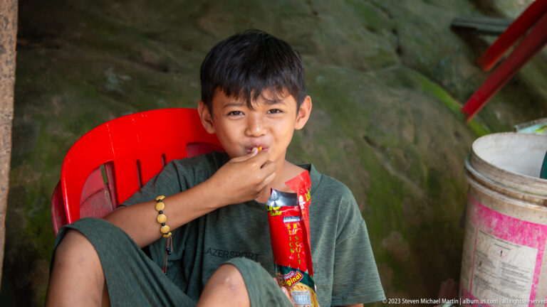 Young boy in a red plastic chair eats from a snack packet and looks at the camera.
