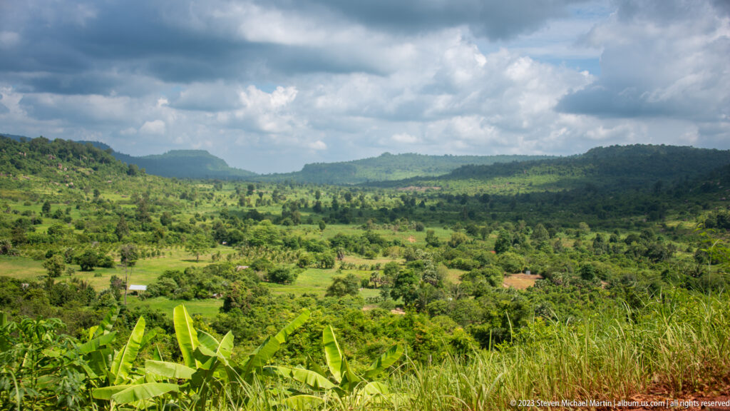 Green cultivated valley and forested ridges seen from Phnom Kulen beneath a cloudy sky.