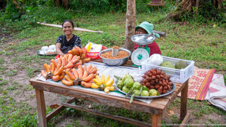 Smiling roadside vendor behind a table of bananas, mixed fruit, baskets, and a hanging scale.