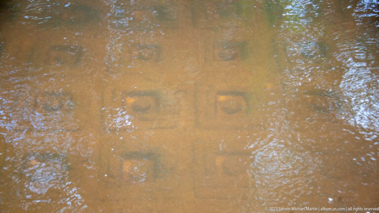 Square and circular stone carvings visible beneath shallow rippling river water.