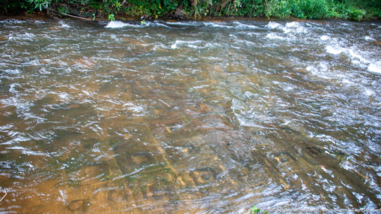 Shallow rapid flowing over rows of circular and geometric riverbed carvings.