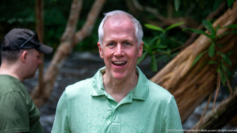 Smiling older man in a pale green shirt with forest and river blurred behind.