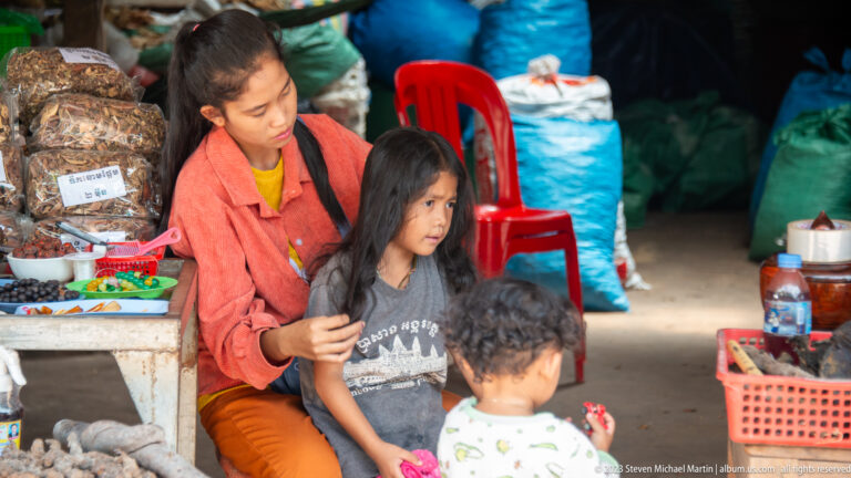 Woman tends a young girl's hair while a small child stands nearby at a market stall.