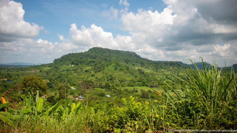 Rounded green hill above fields and scattered homes, framed by tall grass and banana plants.