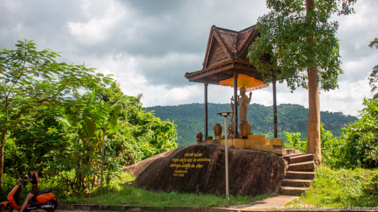 Small roofed shrine with offerings atop a boulder, reached by steps beside a wooded overlook.
