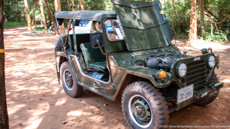 Open-sided vintage Jeep with rugged tires parked on a shaded dirt track in the forest.