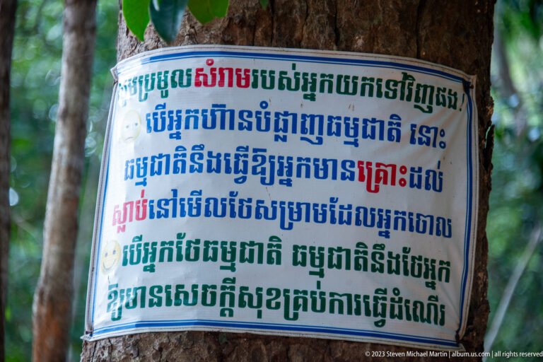 Multicolored Khmer-language notice attached to a tree trunk in a shaded forest setting.