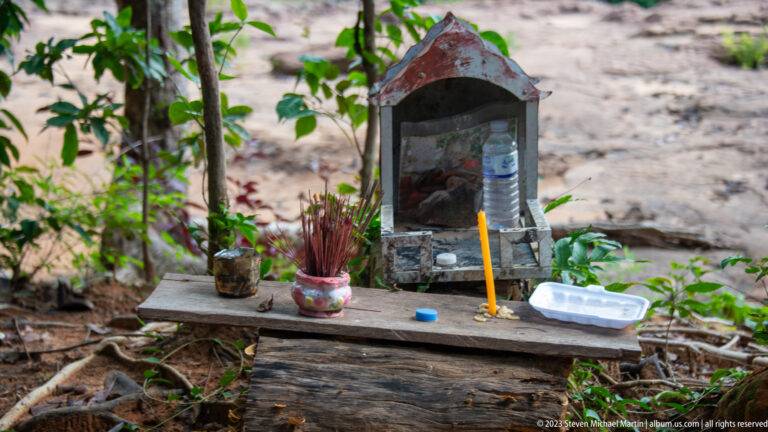 Small weathered shrine with incense, candle, water bottle, and offerings on a wooden bench outdoors.