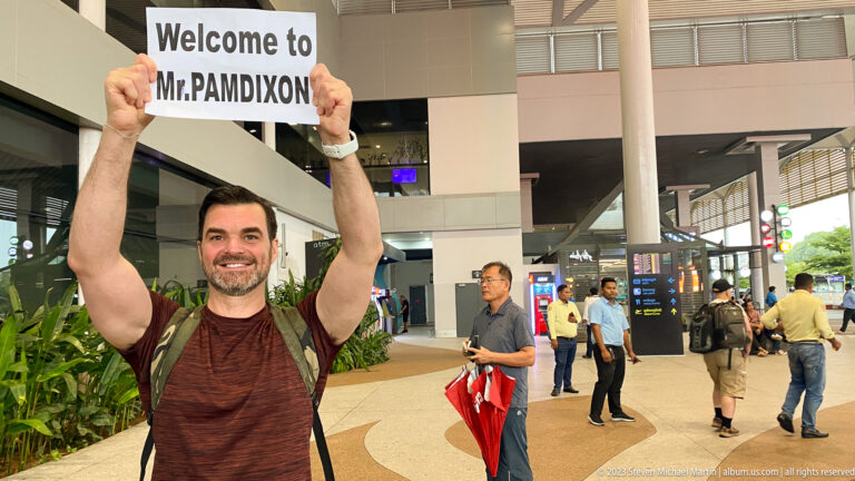 Smiling traveler holds a Welcome to Phnom Penh sign above his head inside a busy airport terminal.