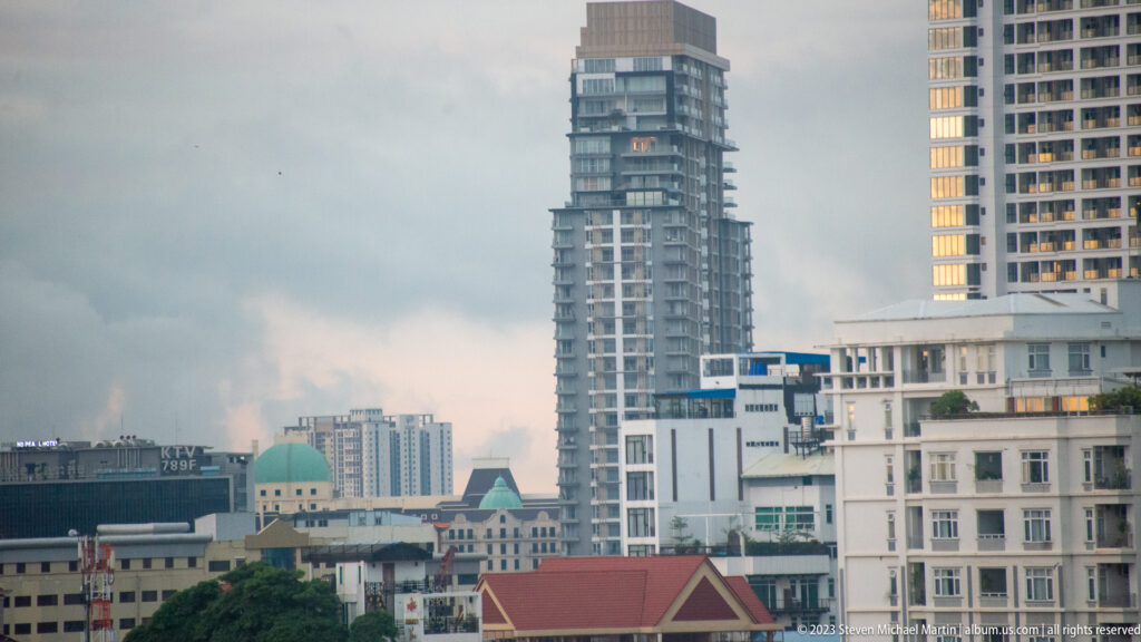 Mixed Phnom Penh skyline with glass towers, unfinished high-rise construction, white buildings, and red roofs.