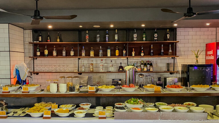 Hotel buffet counter with bowls of salads, fruit, sauces, condiments, and bottles arranged on wall shelves.