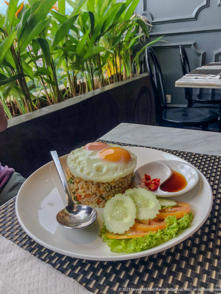 Plate of fried rice topped with a fried egg, cucumber, tomato, lettuce, and sauce at an outdoor table.