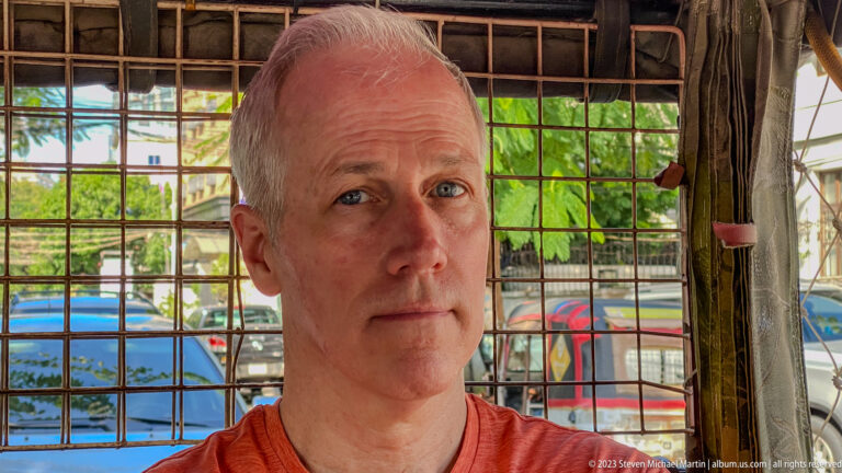 Close portrait of a gray-haired man seated behind metal grillework inside an open vehicle.