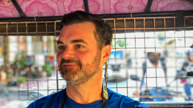 Bearded man in a blue shirt seated behind a metal grille inside an open vehicle.