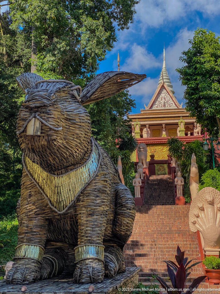 Large seated woven animal sculpture beside Wat Phnom’s brick staircase and temple roof.