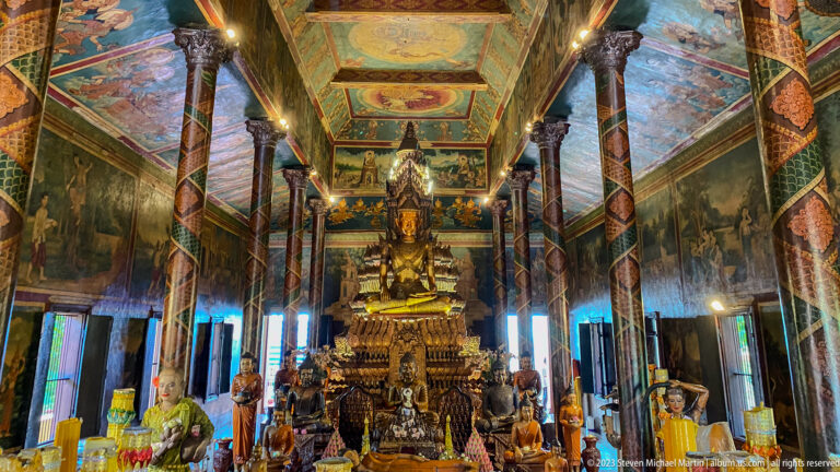 Long symmetrical Wat Phnom interior with painted columns, wall murals, ceiling panels, and central Buddhist altar.