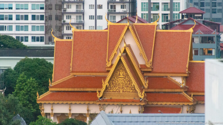 Red-tiled Khmer-style temple roof with gilded gable decoration against modern city buildings.