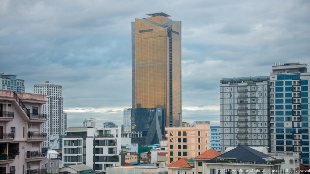 Tall golden high-rise with recessed crown rising above lower apartment blocks and red-roofed buildings.