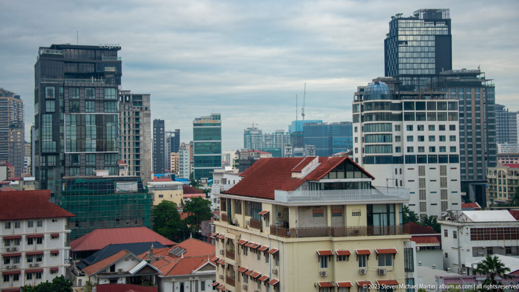 Modern Phnom Penh towers behind older white buildings and red tile roofs under a cloudy sky.